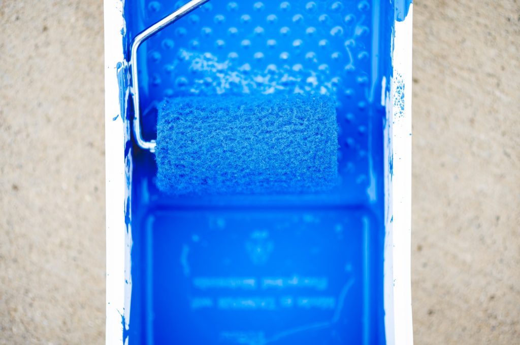 Top-down view of a textured paint roller soaked in vibrant blue pool deck coating sitting in a matching blue paint tray.