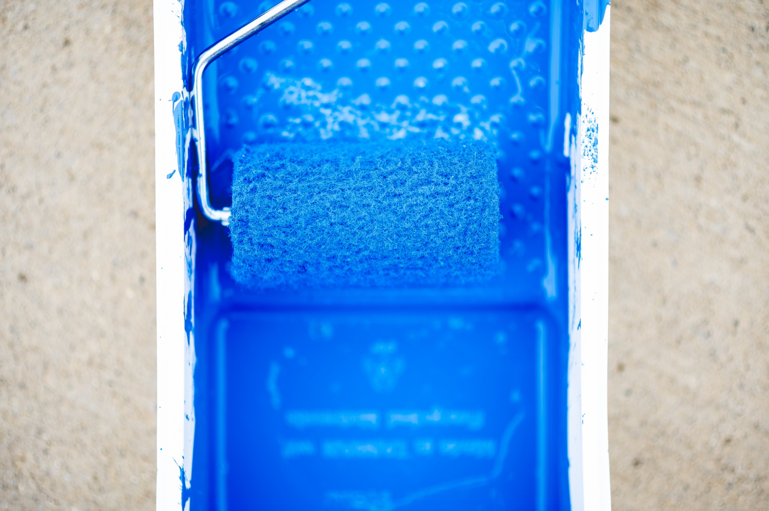 Top-down view of a textured paint roller soaked in vibrant blue pool deck coating sitting in a matching blue paint tray.