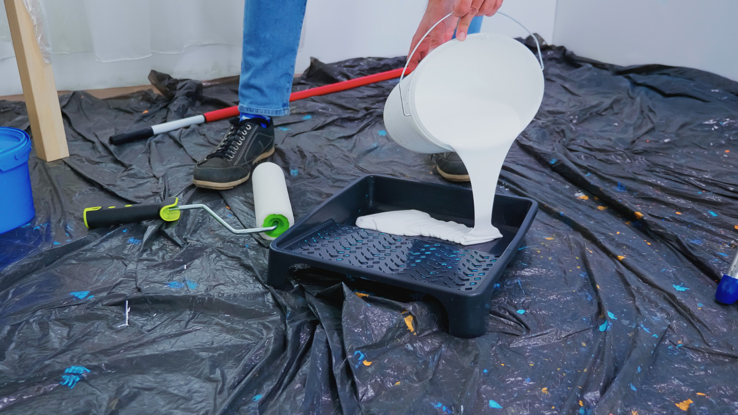 A person pouring thick white liquid rubber coating from a white bucket into a black paint tray on a protected indoor floor surface.