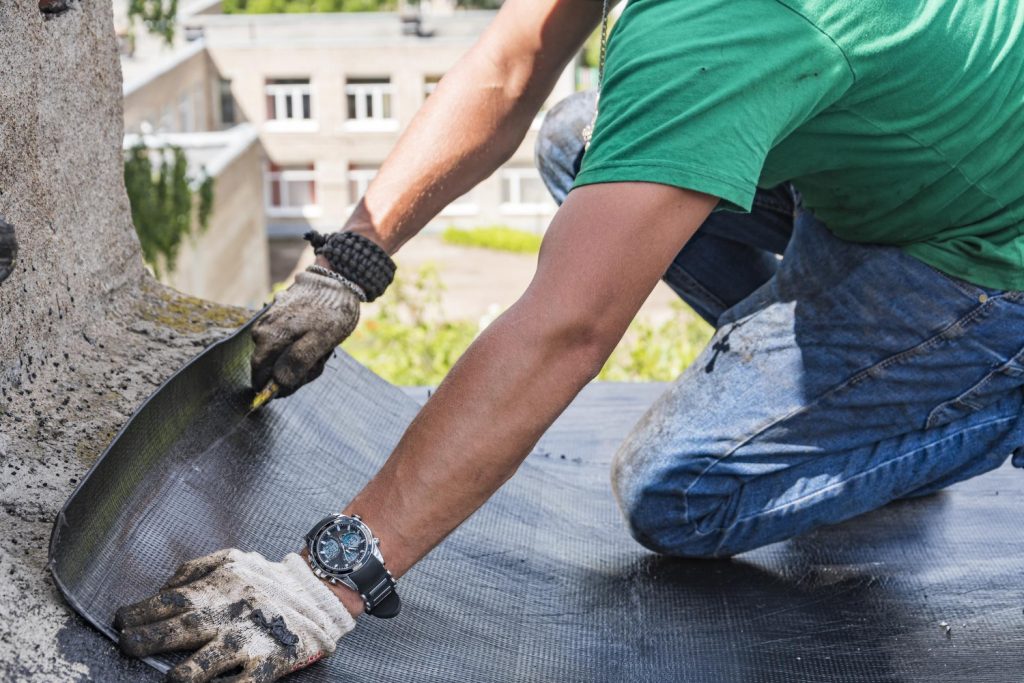 A roofing professional wearing protective gloves installs a durable black rubber membrane coating along a roof edge and parapet wall.