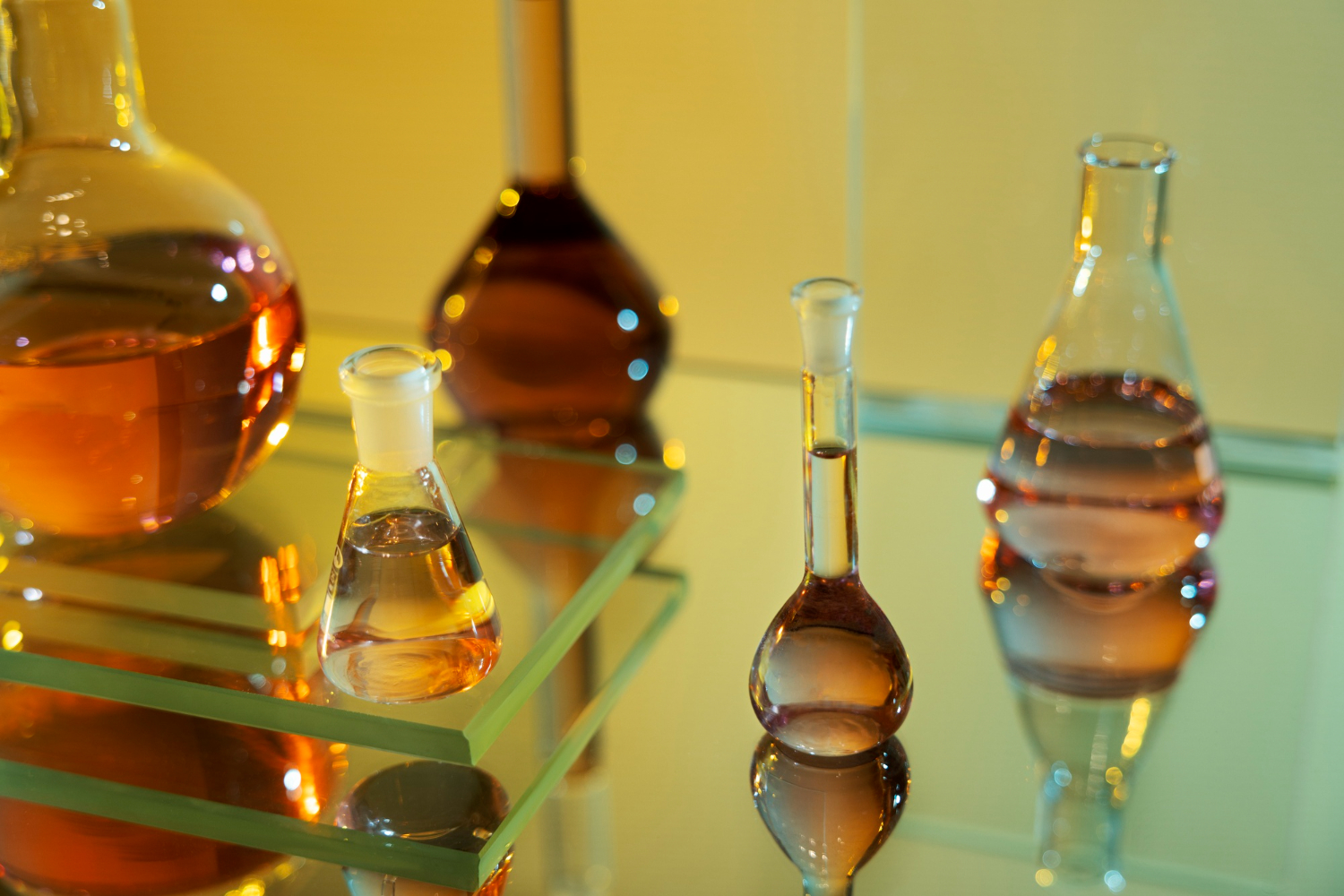 Various glass laboratory flasks and beakers filled with amber and clear surfactant chemical liquids arranged on a reflective glass surface.