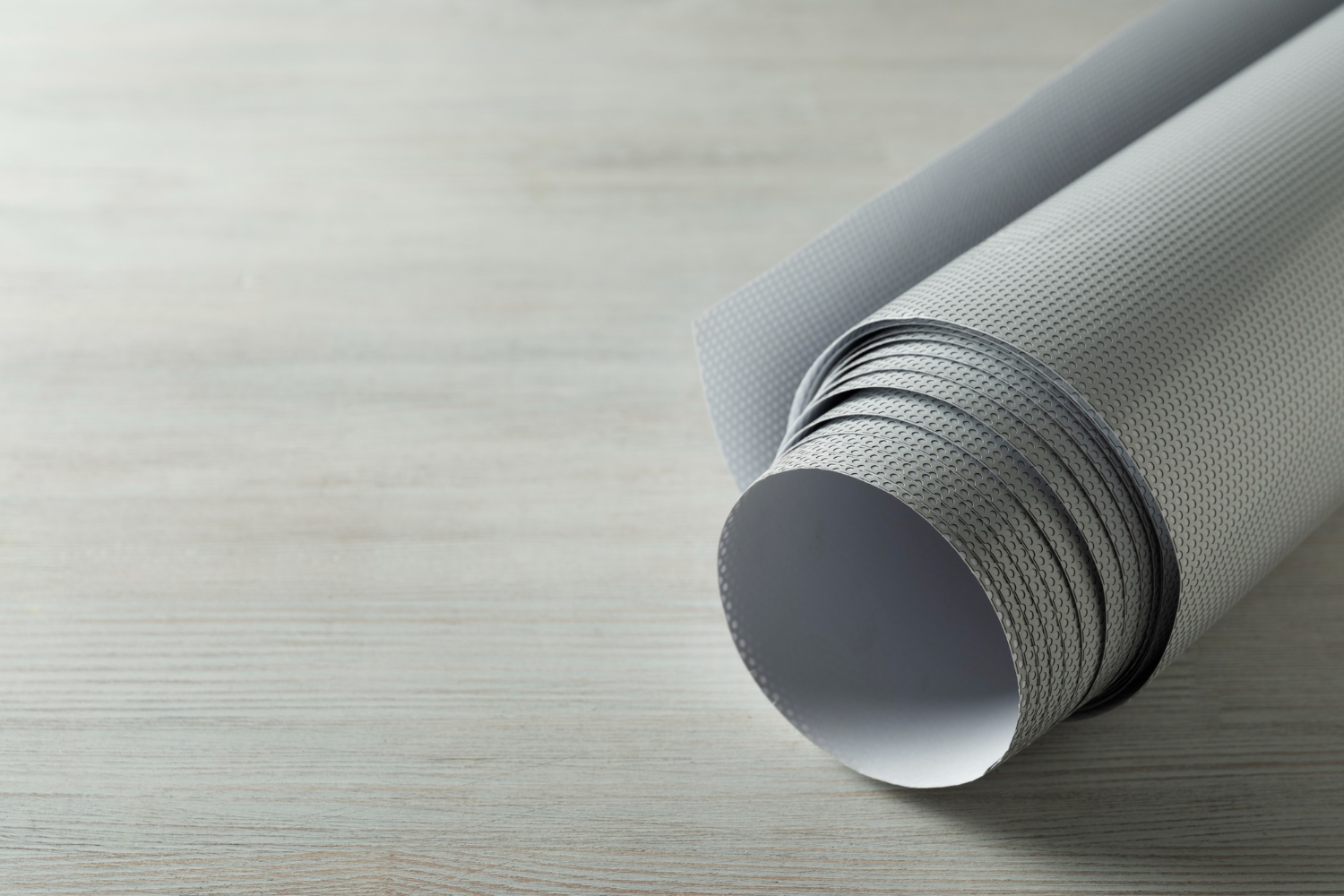 A roll of grey TPO roofing membrane showing its reinforced textured surface and white underside, resting on a light wood background.