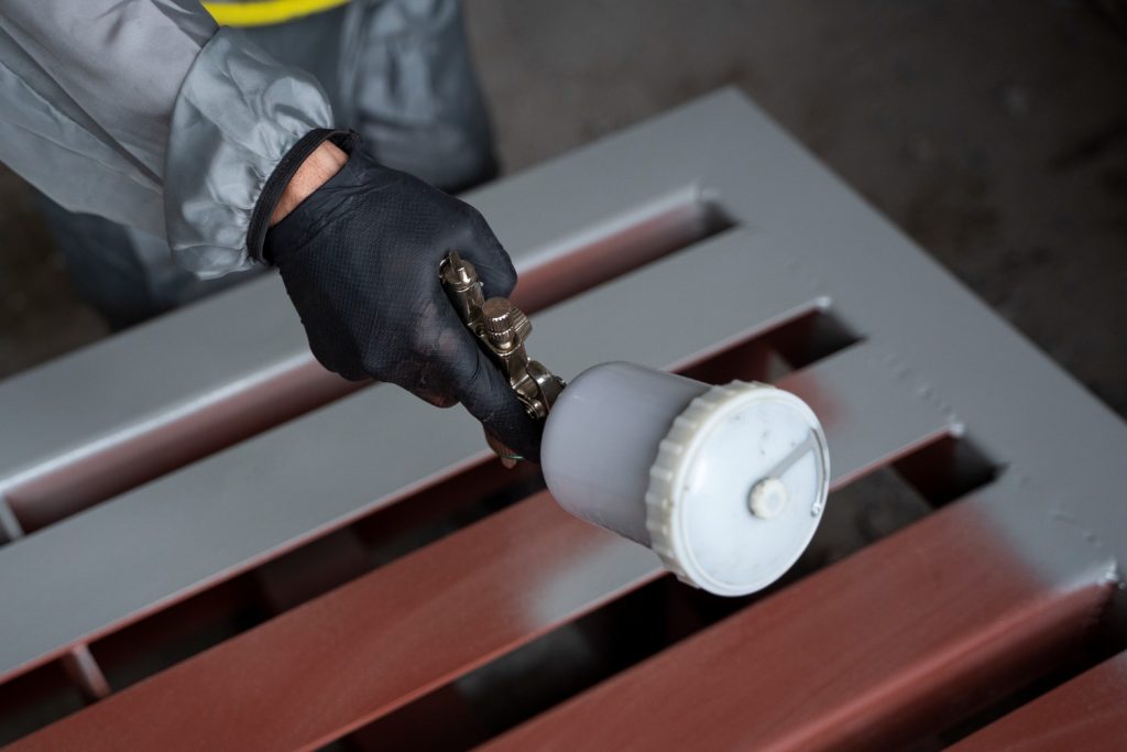 Close-up of a technician in protective gear and black gloves using a precision spray gun to apply a silver PVD coating to a red metal industrial component.