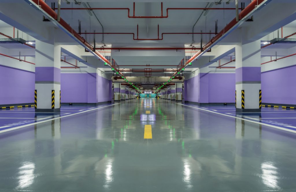 A symmetrical view of a large, high-gloss gray polyaspartic floor in a modern commercial parking garage with purple walls, white pillars, and red overhead piping.